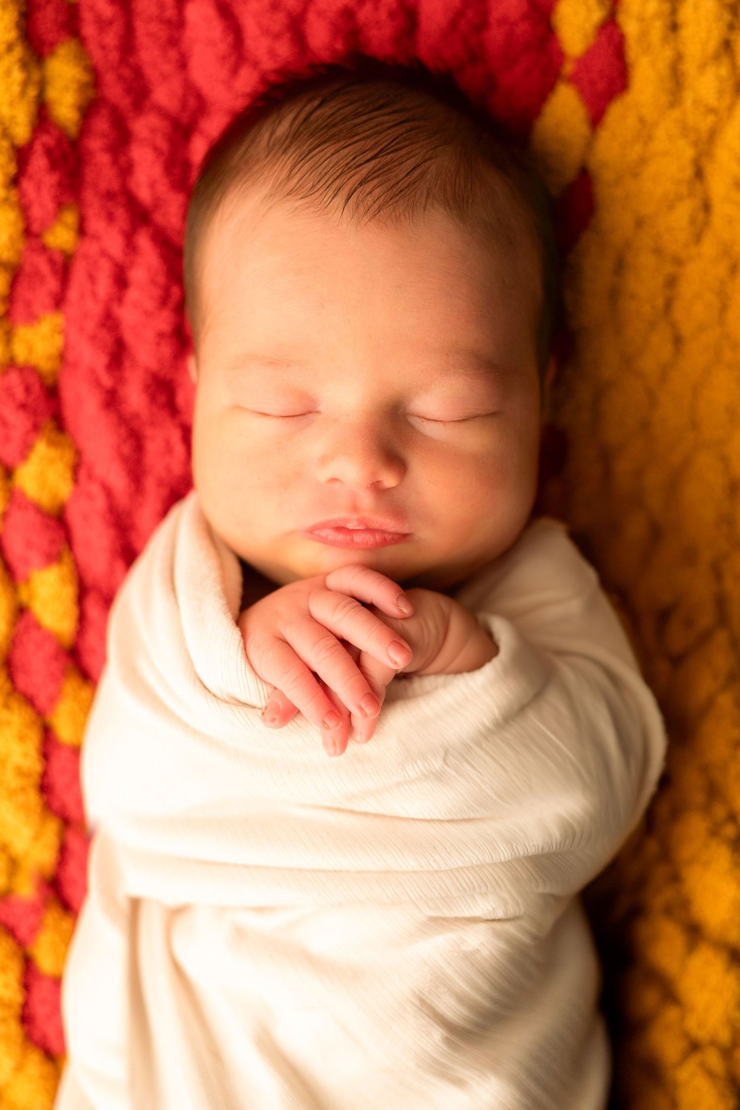 A sleeping newborn baby swaddled in white fabric rests peacefully against a vibrant red and yellow knitted blanket background.