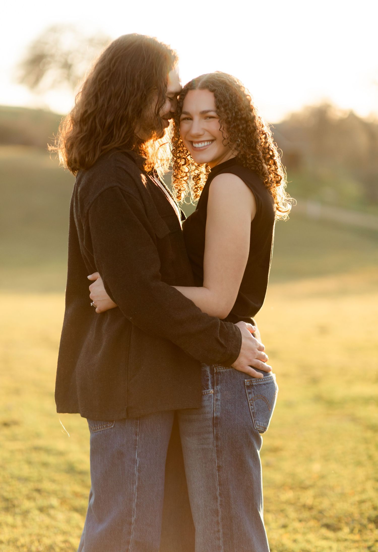 Romantic couple embraces in a golden sunset field while wearing casual attire.