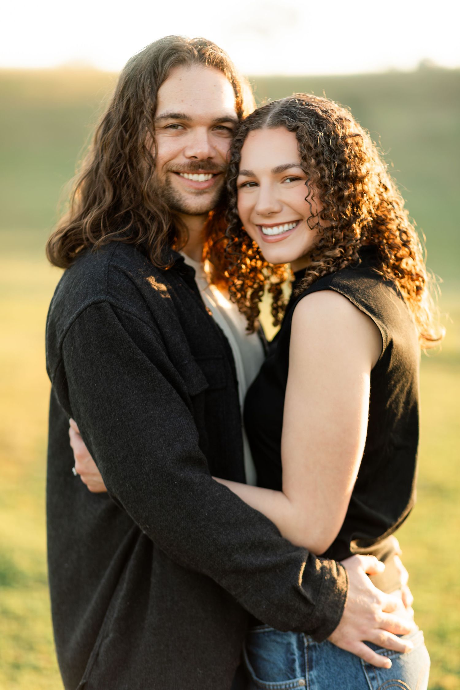 A couple embraces and laughs together in a golden sunset field with warm backlighting.