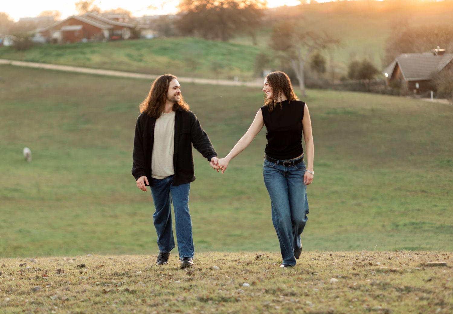 A couple walks hand in hand through a grassy field during sunset.