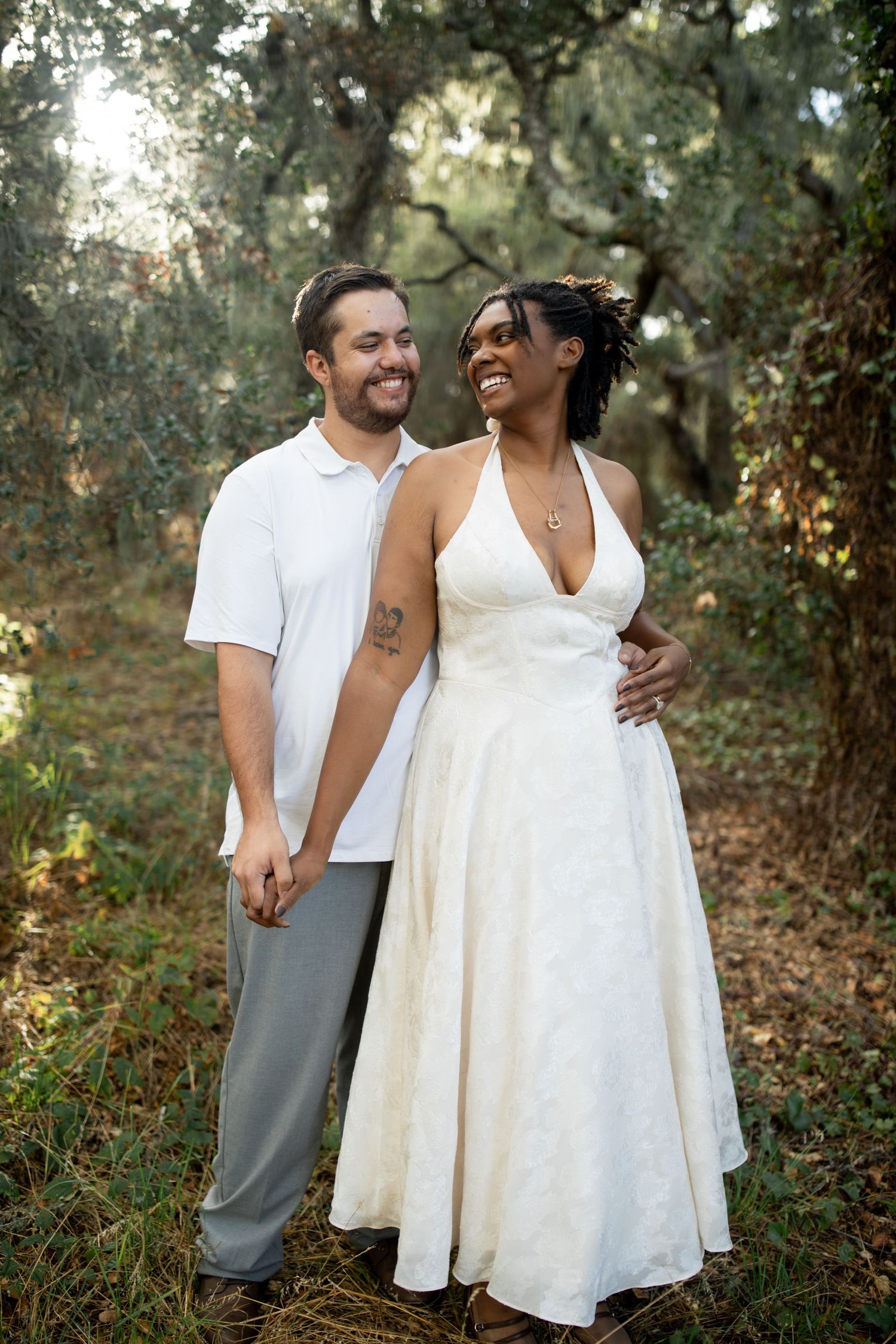 A couple shares intimate moments in a wooded outdoor setting wearing white and grey casual attire.