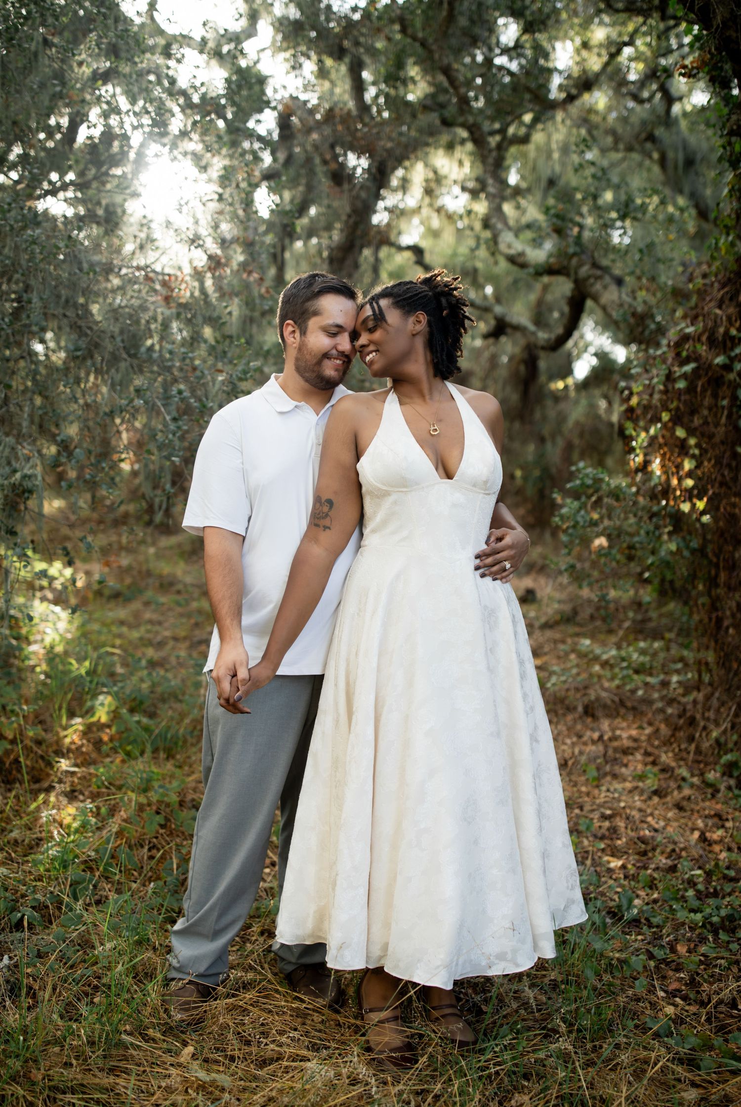 A couple in white and gray attire share intimate moments in a wooded outdoor setting during a romantic photoshoot.