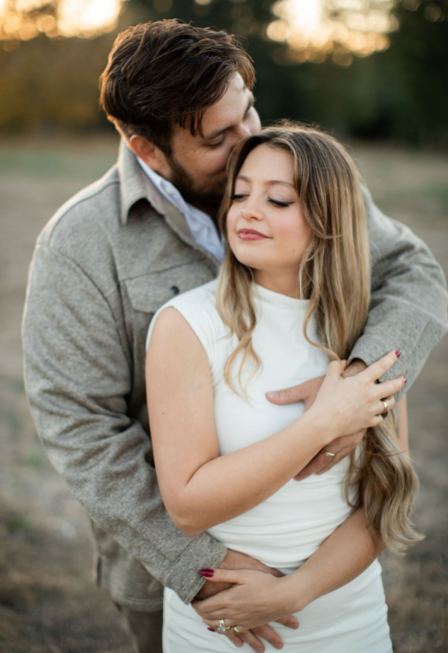A couple embraces intimately during a golden hour outdoor photo session wearing white and gray clothing.