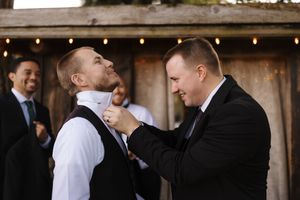 A groomsman helps adjust another's tie at an outdoor wedding venue with string lights and wooden walls in the background.