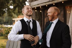 Wedding party members in black suits and white shirts laugh together during outdoor photo session at sunset.