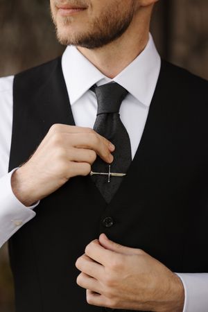 Close-up view of someone adjusting a black tie with a tie clip while wearing a formal black vest and white dress shirt.