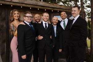 A wedding party in formal attire poses together outdoors with black suits and pink dresses against a rustic backdrop.