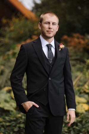 A groom in a classic black suit poses outdoors against a natural autumn backdrop with blurred foliage.