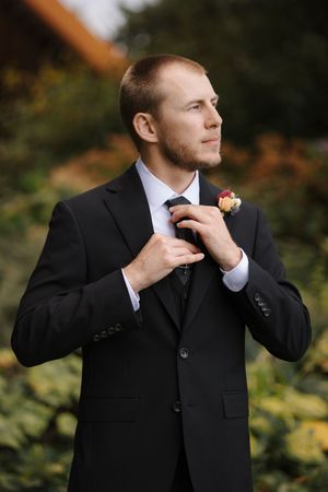 A groom in a black suit adjusts his tie while standing outdoors with autumn foliage in the background.