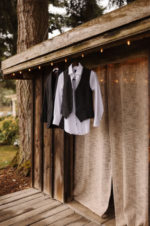 Wedding attire including white shirts and black vests hanging on rustic wooden wall with string lights.