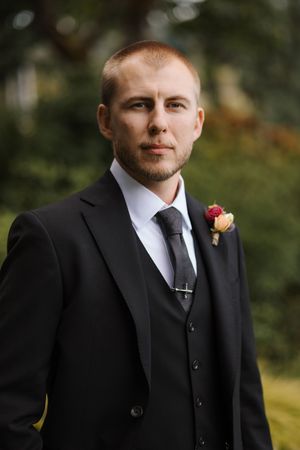 A groom in a black three-piece suit with a pink boutonniere poses for a formal wedding portrait outdoors.