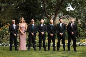 Wedding party in formal attire poses outdoors in a line formation against a garden backdrop.