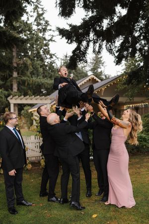 Wedding party members in black suits and pink dresses perform a fun group pose outdoors with string lights in background.