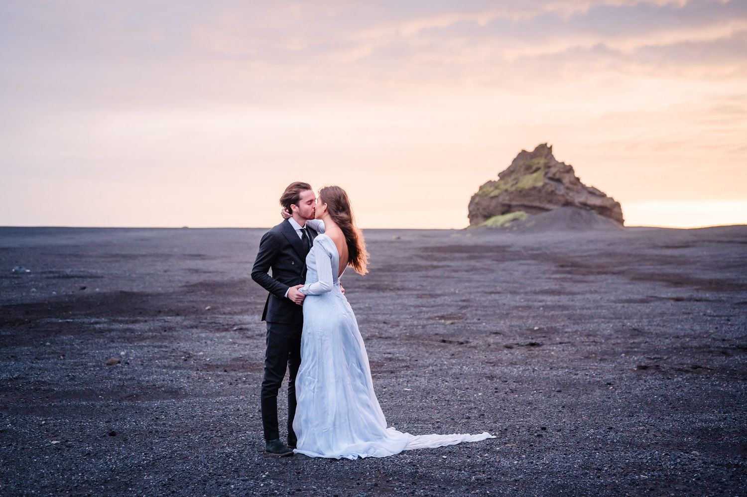 A couple embraces on a dramatic black sand beach at sunset with ocean cliffs in the background.