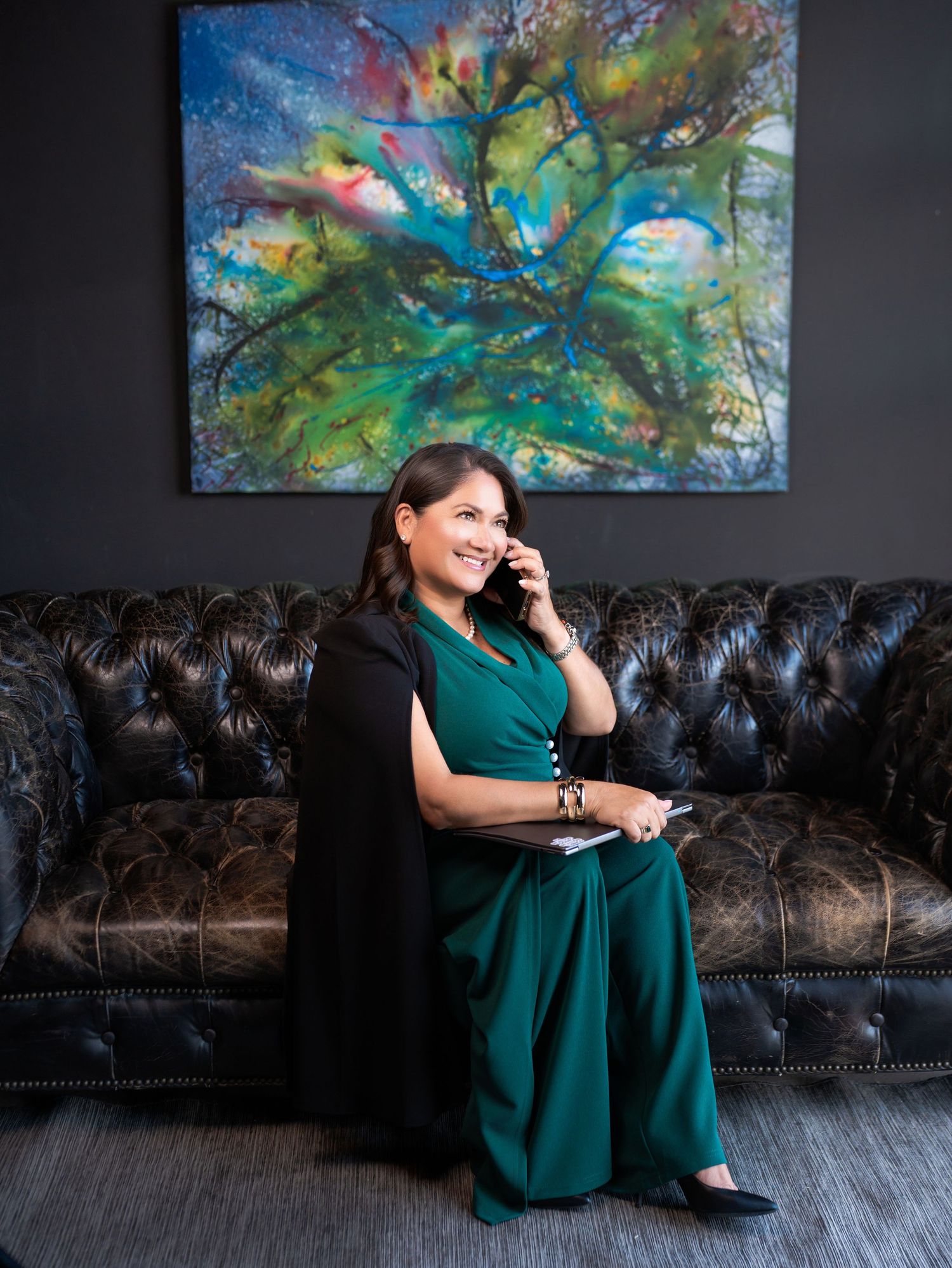 A person in an emerald green dress poses casually on a brown leather couch with colorful abstract artwork displayed on the wall.