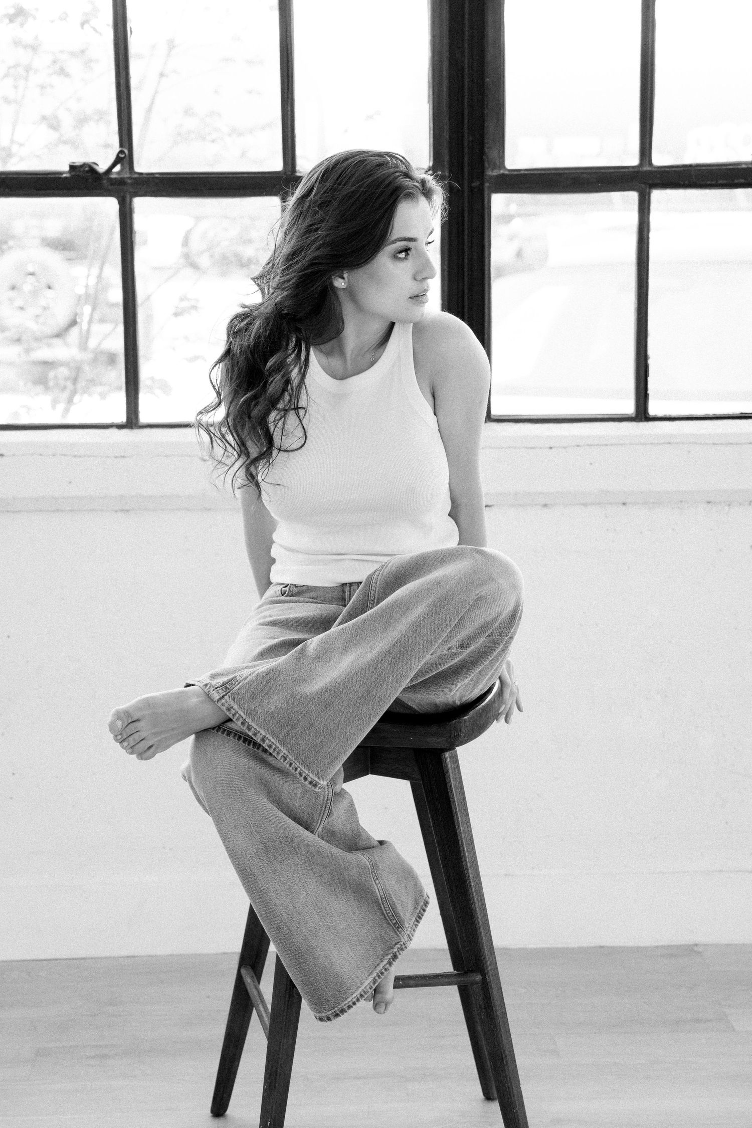 Black and white photo of a woman in casual attire sitting cross-legged on a wooden stool near a window.