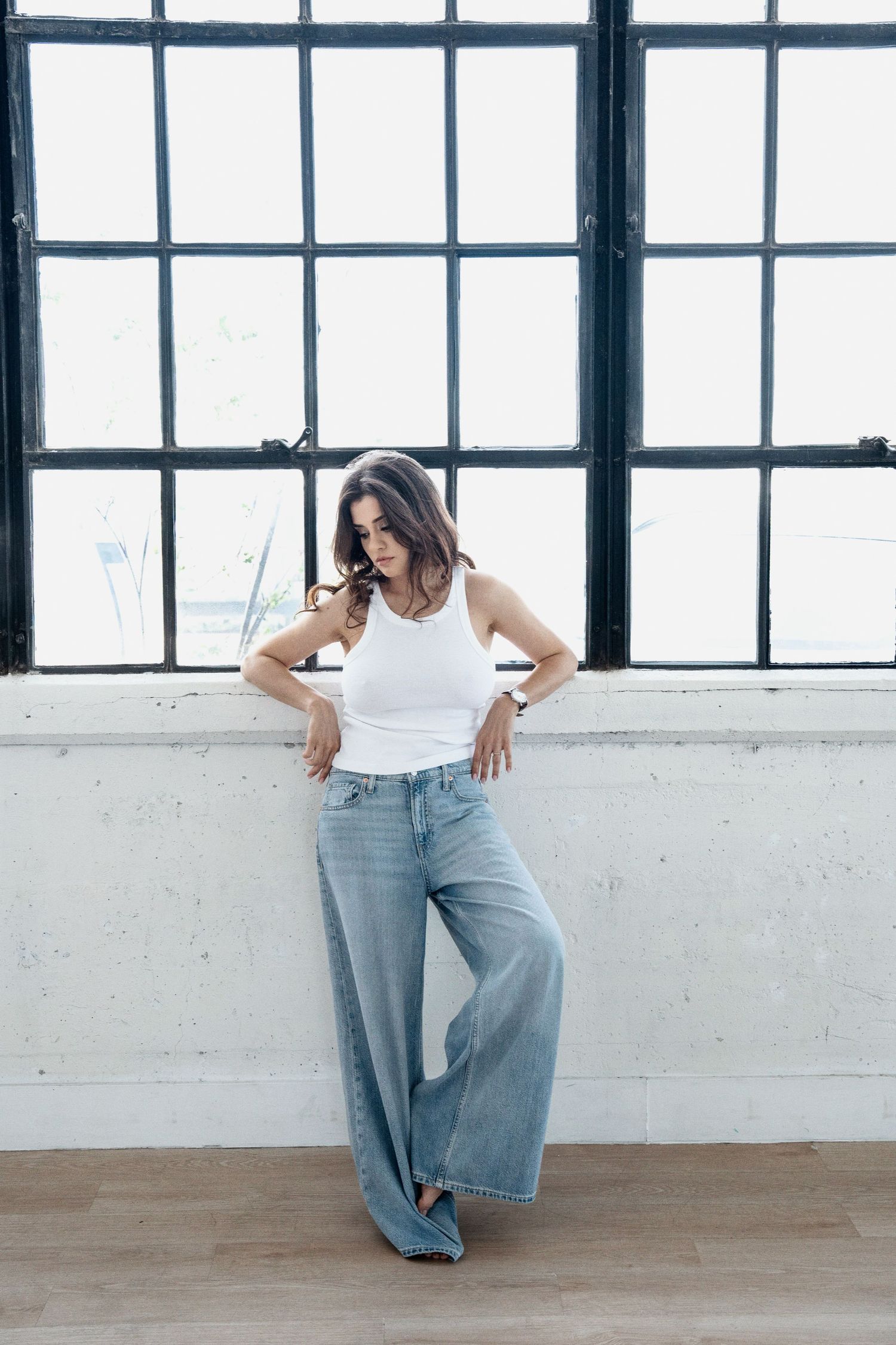 Woman in white tank top and wide leg jeans poses dynamically against industrial windows in studio setting.