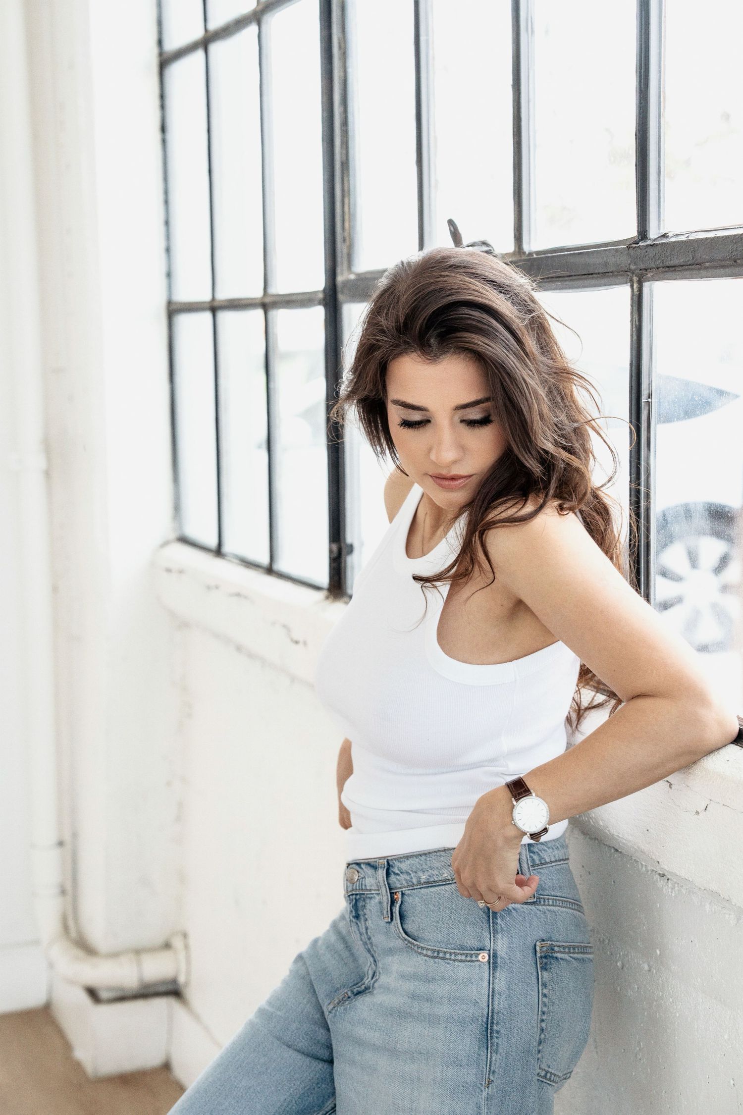 Fashion model in white top and jeans poses casually against bright industrial windows.