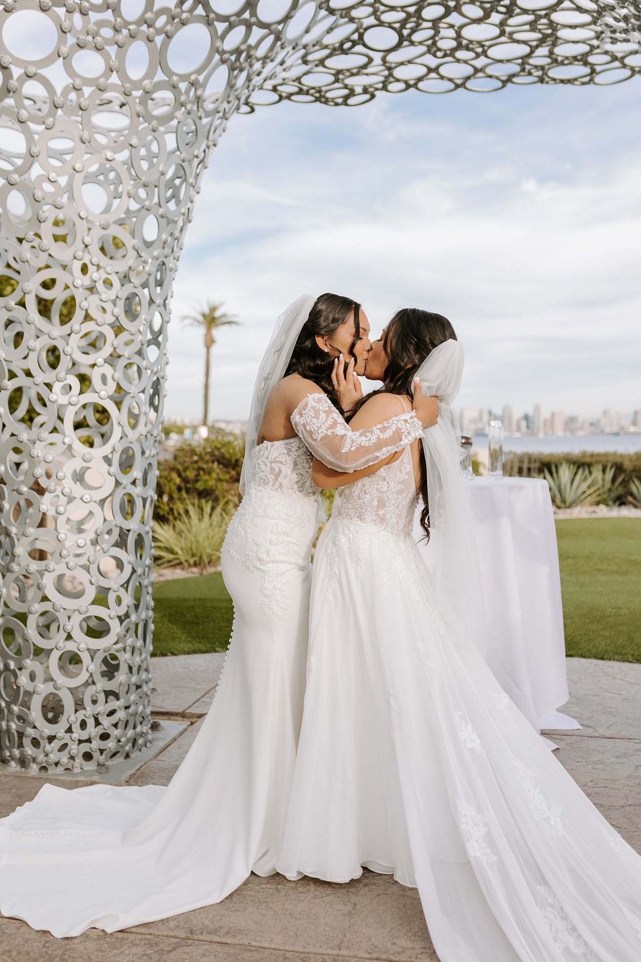 Two brides share their first kiss during their wedding ceremony under an ornate white lattice arch at an outdoor venue.