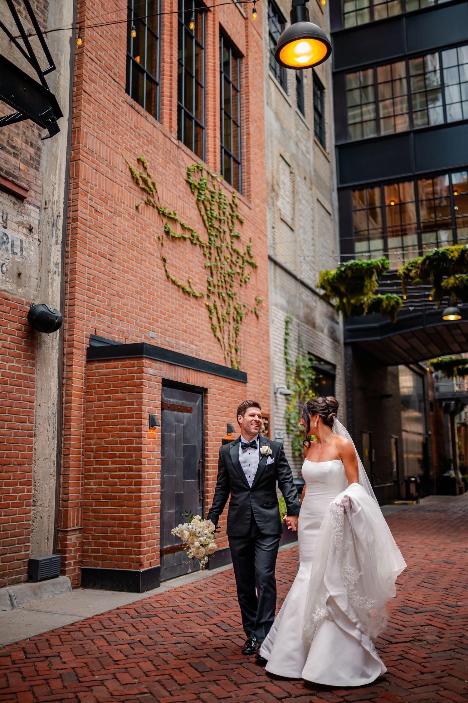 Smiling bride in lace gown sits on cream sofa leaning against groom in black suit holding white rose bouquet.