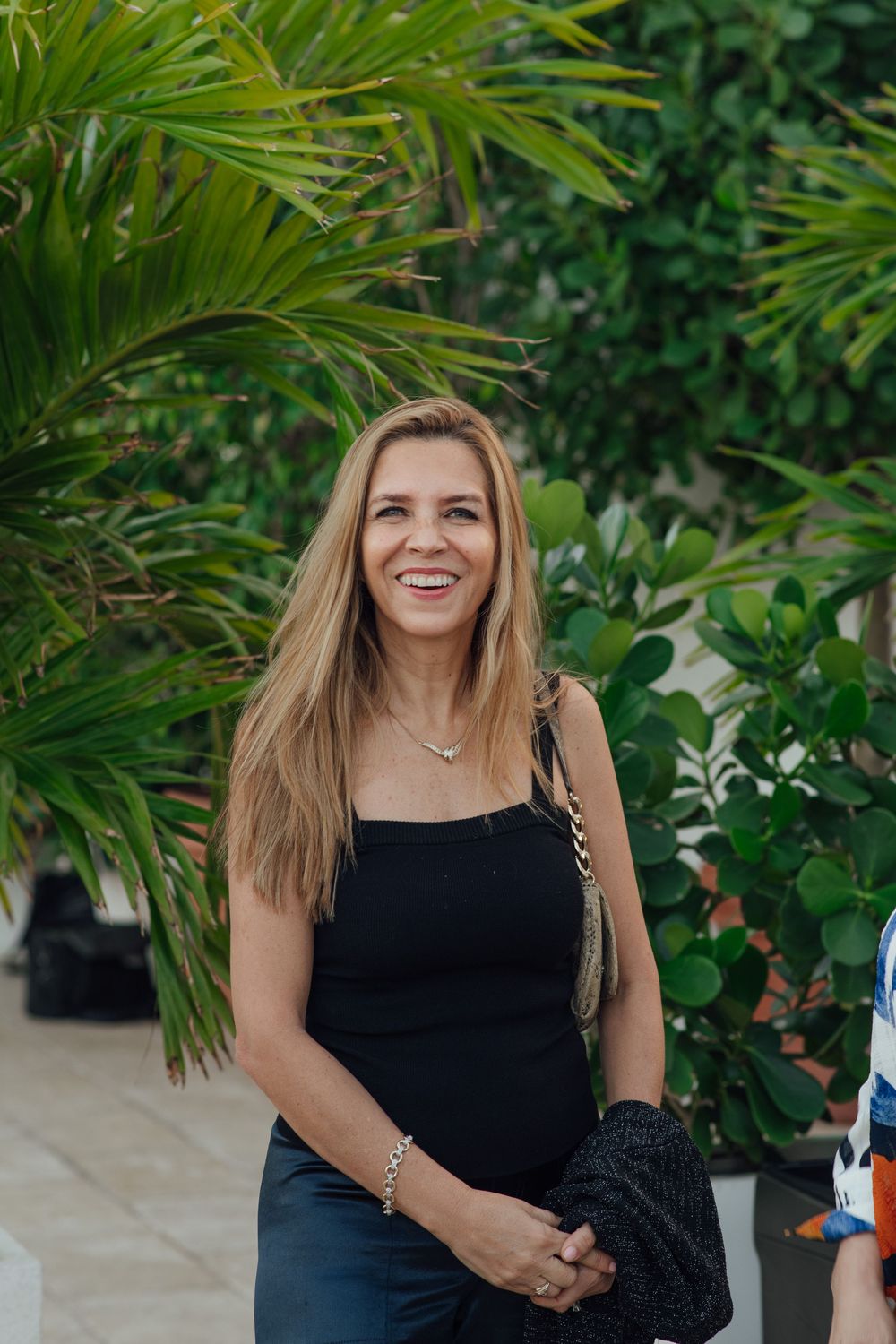 Smiling blonde woman in black outfit holding a handbag at a tropical outdoor event.