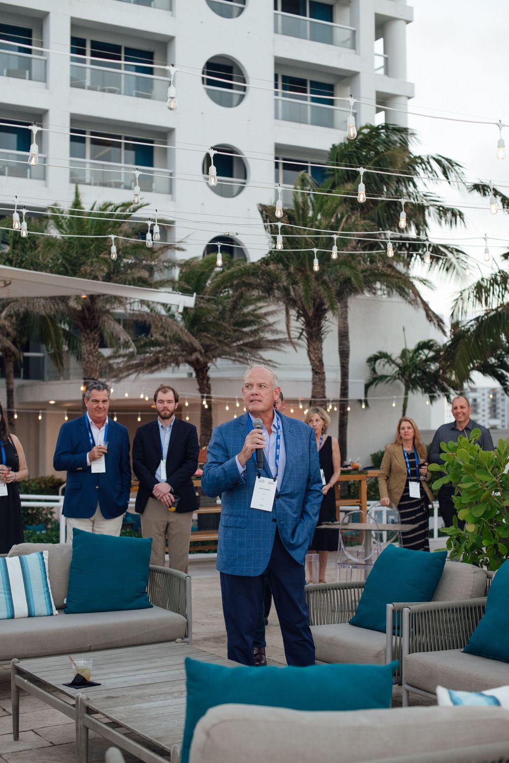 Man in blue suit speaking into microphone at outdoor conference event with hotel backdrop.