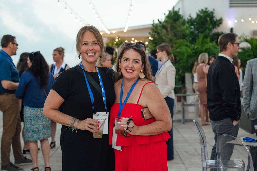 Two smiling women holding drinks and posing together at an outdoor evening networking event.
