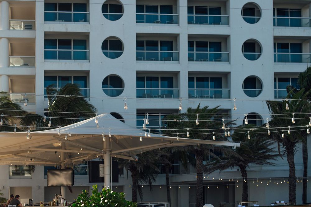 White beachfront hotel with circular windows, palm trees, and string lights over an outdoor terrace.