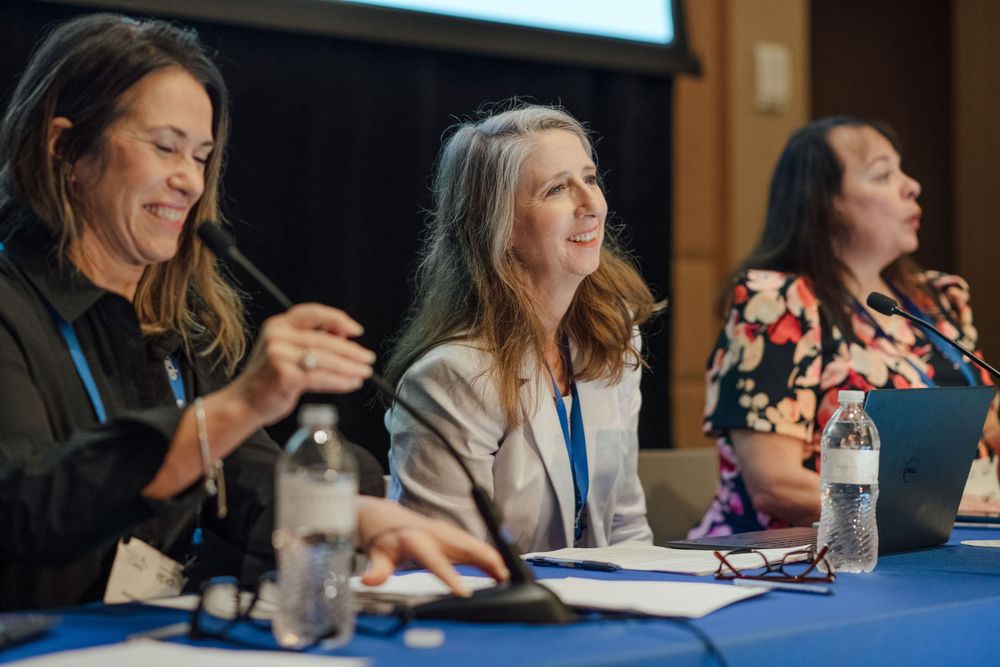 Three women smiling and laughing together at blue conference table panel discussion.