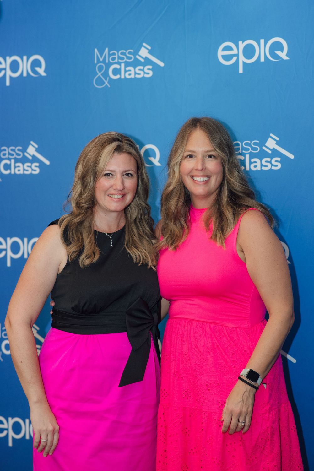 Two women in pink and black outfits posing at Epiq Mass & Class event backdrop.