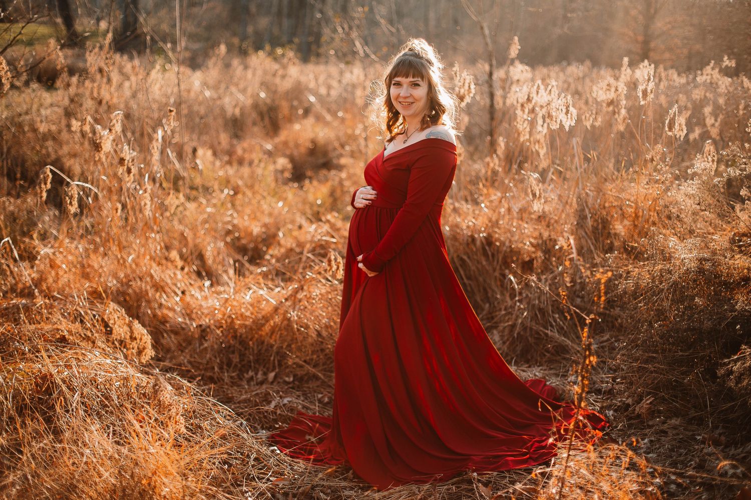 Pregnant woman in flowing red gown poses in golden autumn field during maternity photoshoot.