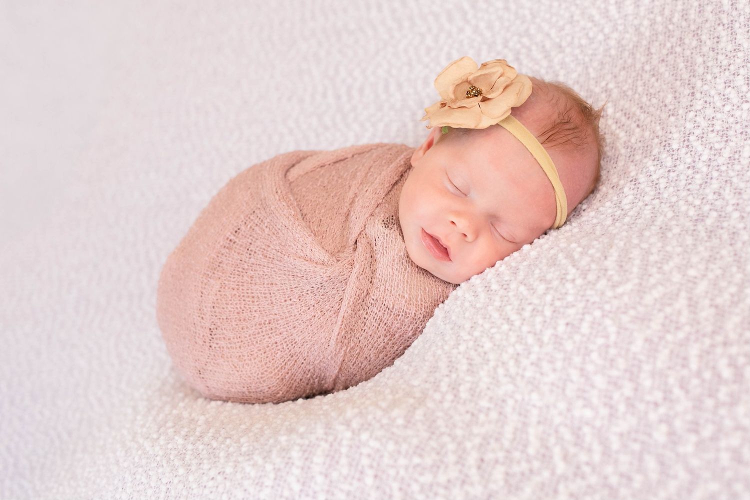 Sleeping newborn baby swaddled in pink wrap with floral headband on white blanket.