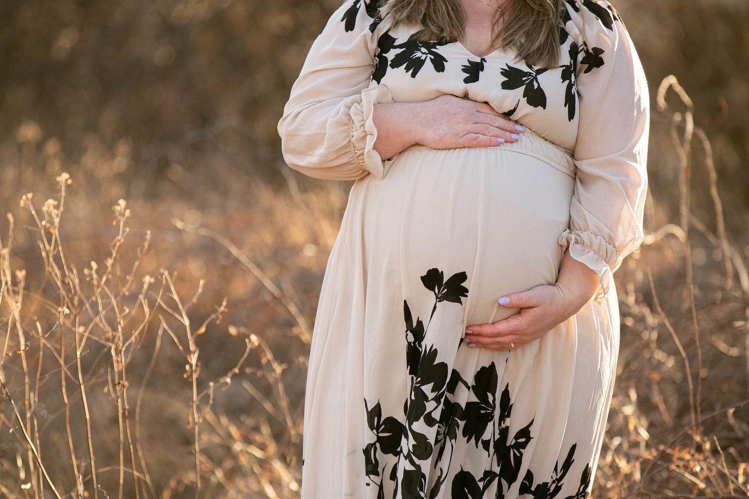 Pregnant woman in floral cream dress cradling baby bump in golden field during maternity photo shoot.