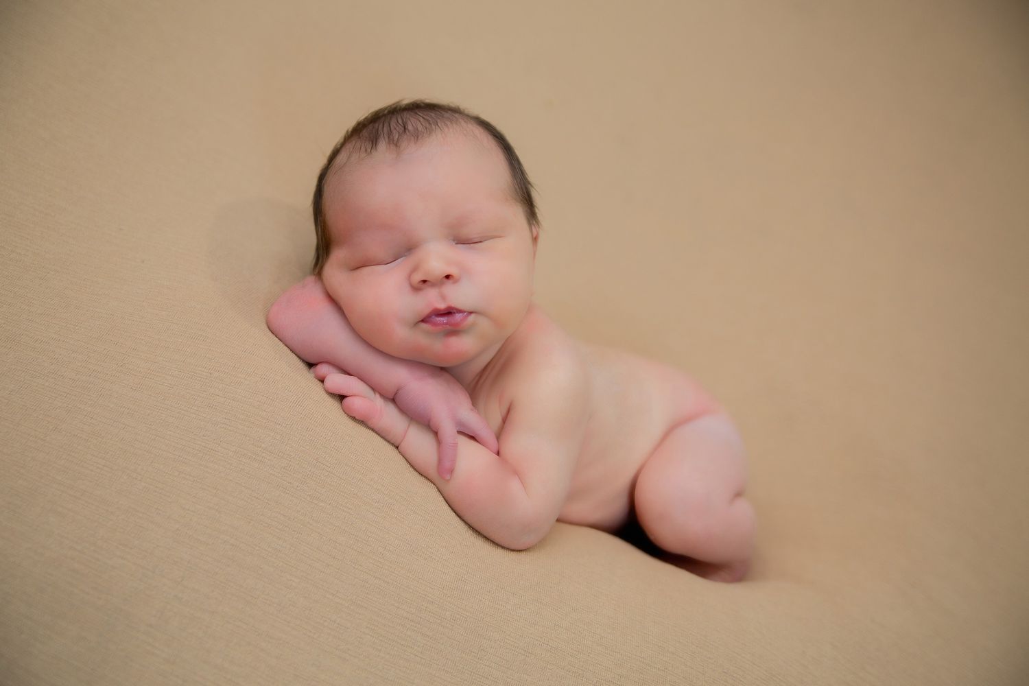 Peaceful newborn baby sleeping curled up on a soft beige blanket in a classic posed newborn photography style.