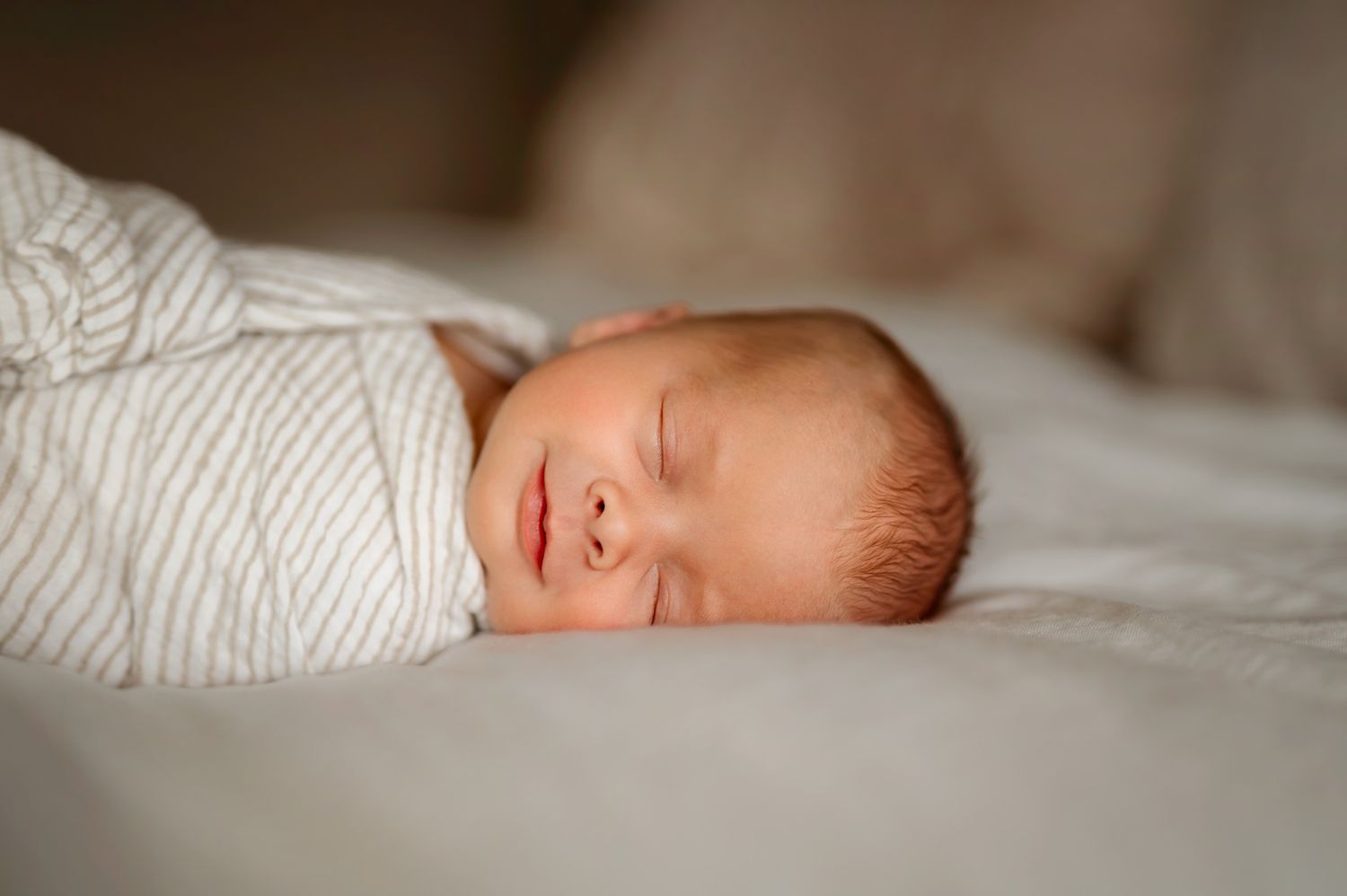Peaceful newborn baby swaddled in white striped blanket sleeping on soft white bedding.