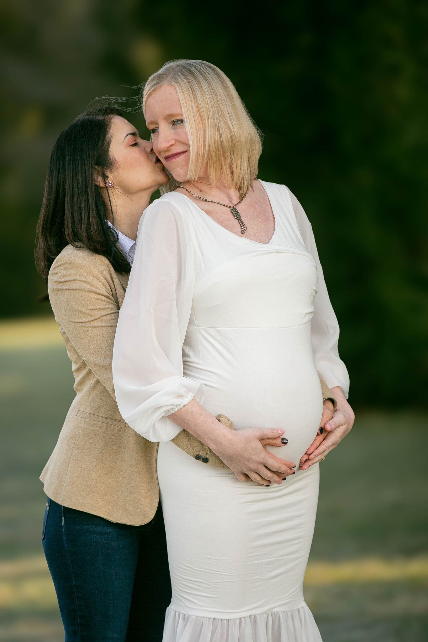 Two women outdoors, one kissing the cheek of a smiling pregnant woman in a white dress.