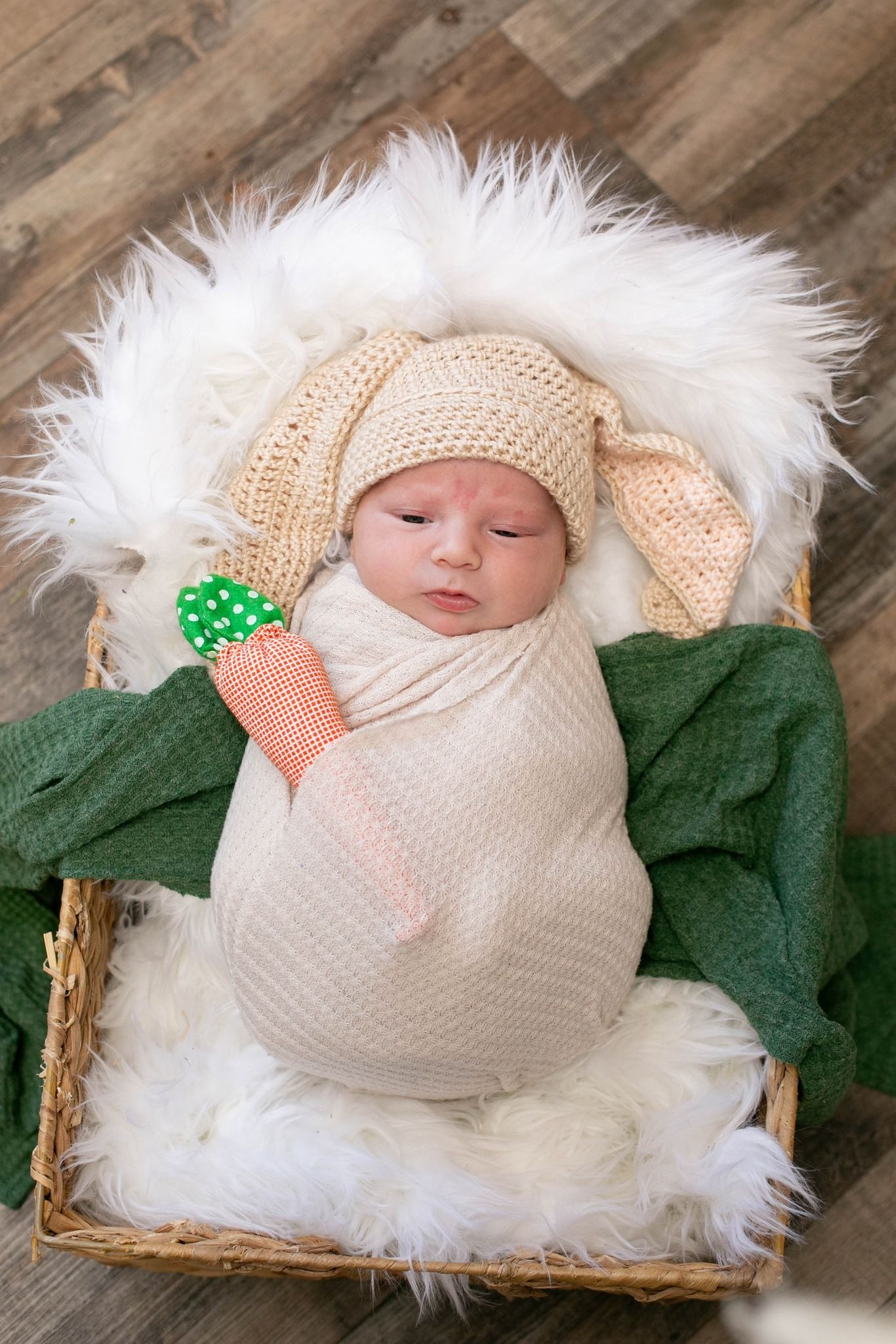 Newborn baby swaddled in cream wrap, wearing bunny ear hat, holding toy carrot, resting in wicker basket with white fur.