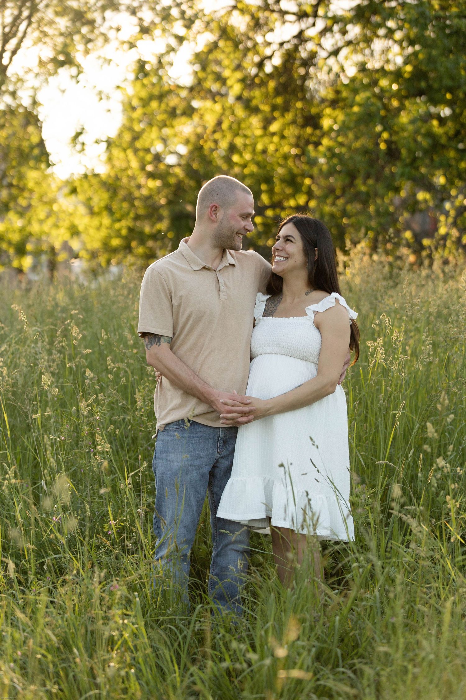 Happy pregnant couple embracing in a sunny green meadow, woman in white dress smiling at partner.