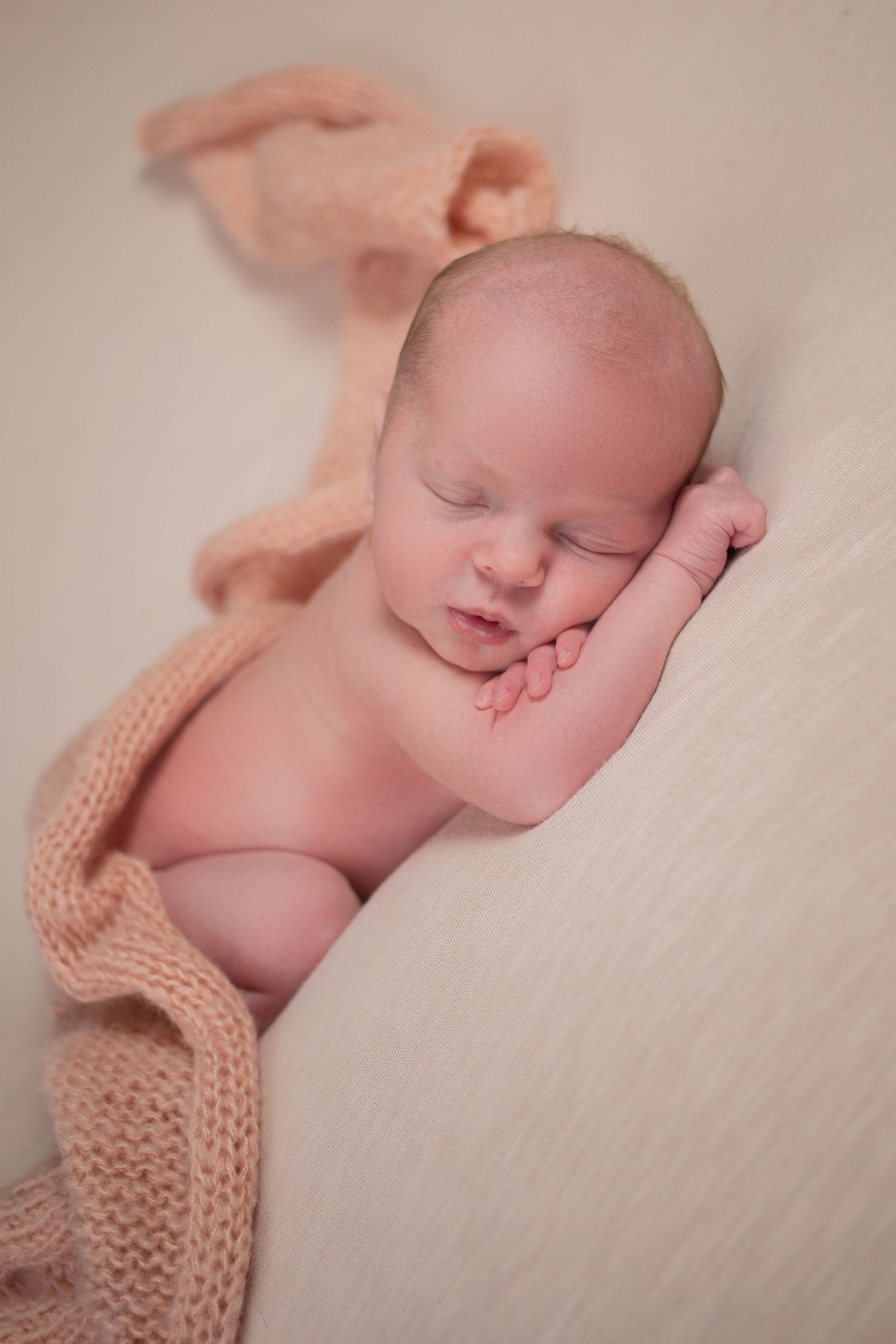 Peaceful newborn baby sleeping on a soft cream blanket, wrapped in a cozy peach knit wrap, hands tucked under cheek.