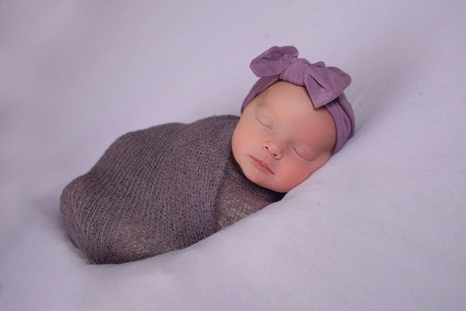 Sleeping newborn baby swaddled in gray knit wrap wearing purple bow headband on lavender background.