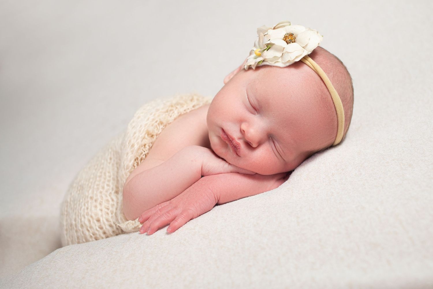 Peacefully sleeping newborn baby girl wrapped in cream knit swaddle with floral headband on white backdrop.