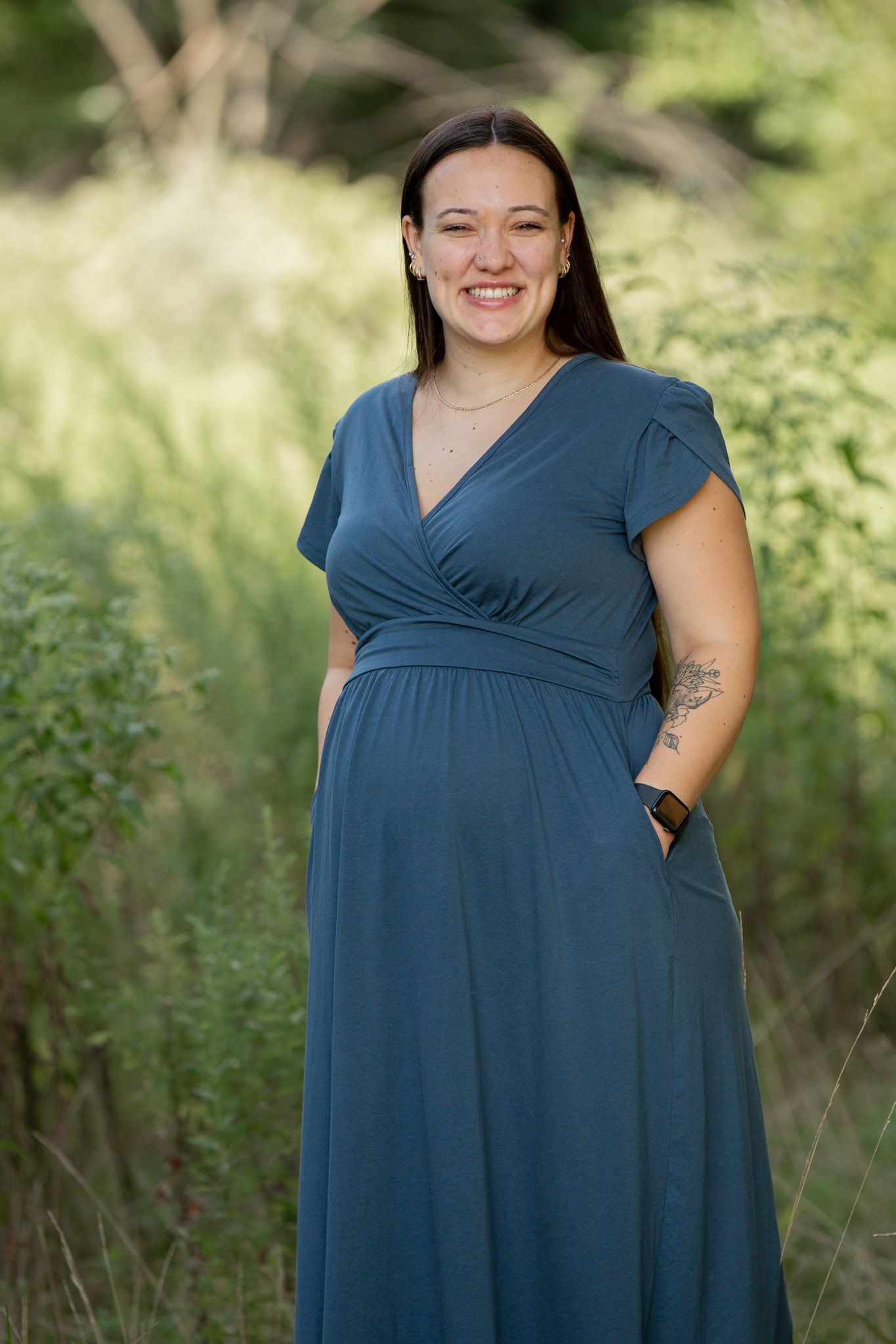 Smiling woman in teal maxi dress standing outdoors among lush green foliage in a natural setting.