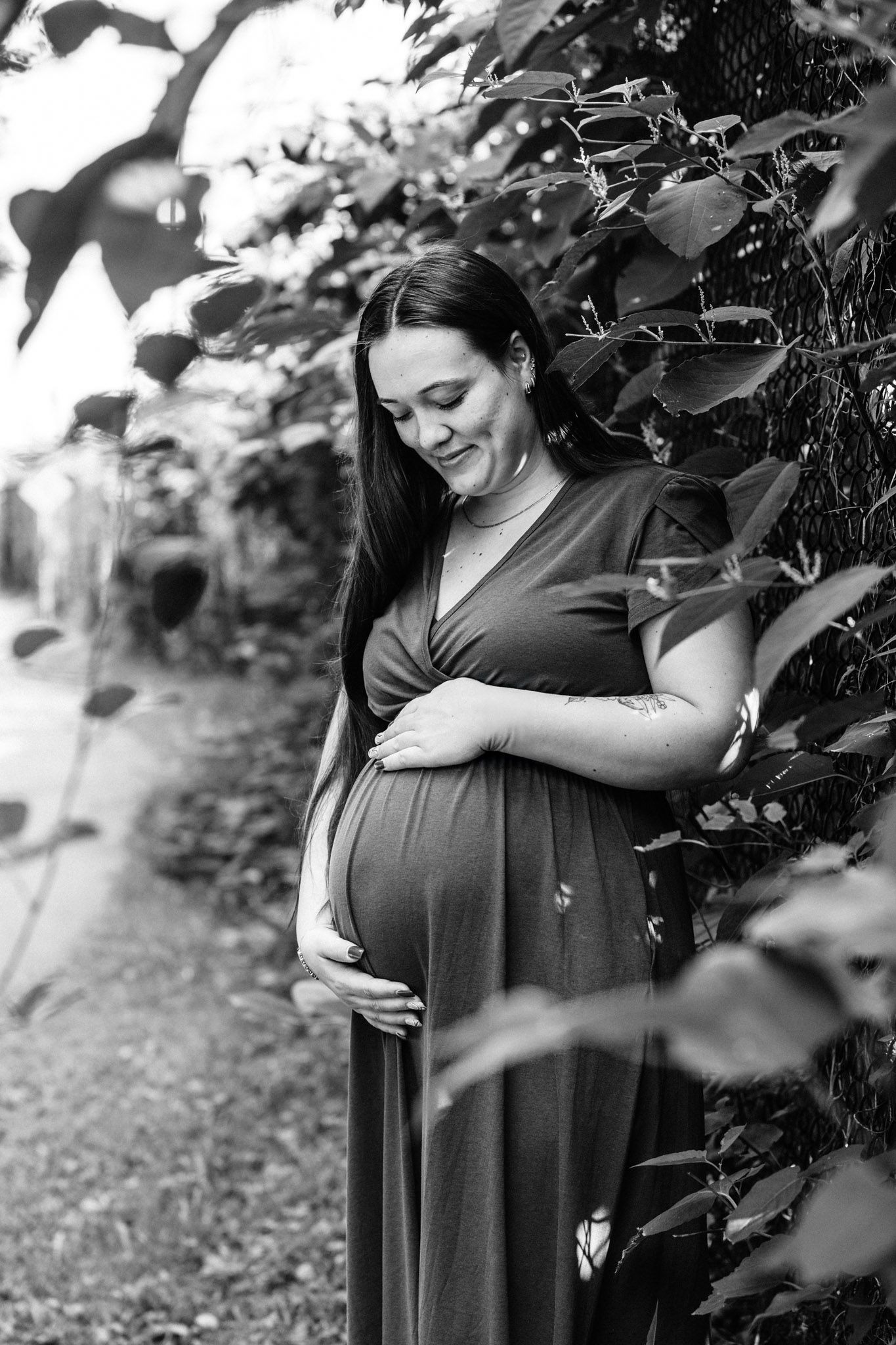 Black and white photo of a pregnant woman in a maxi dress cradling her baby bump outdoors among lush green leaves.