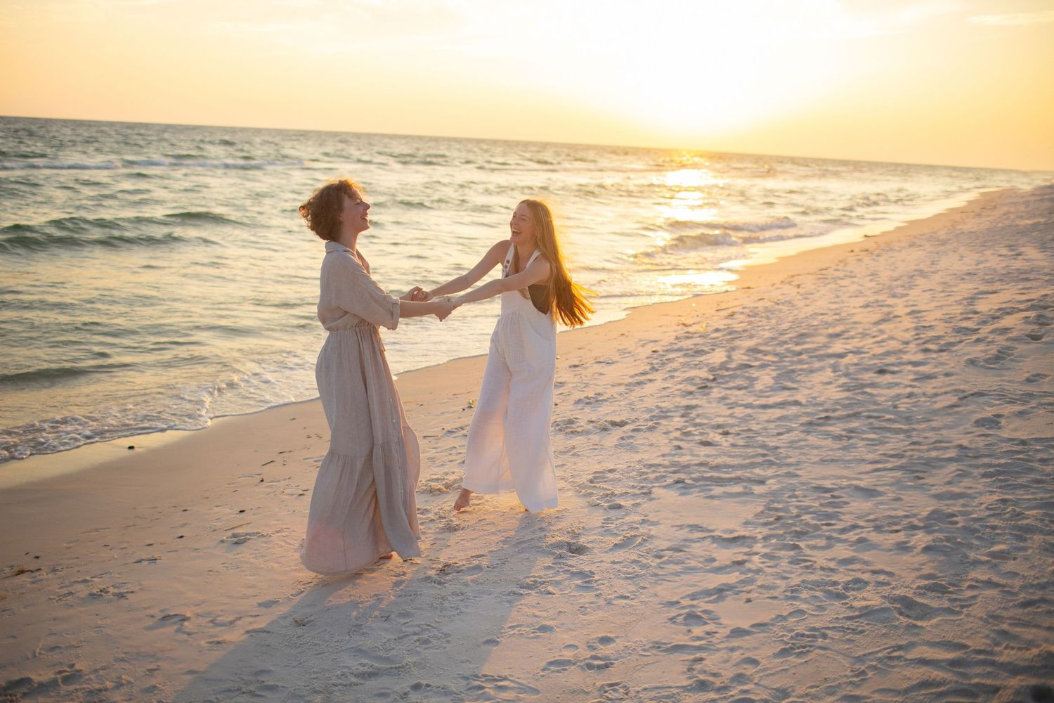 Two people in white dresses walk hand in hand along a sandy beach at sunset with ocean waves in the background.