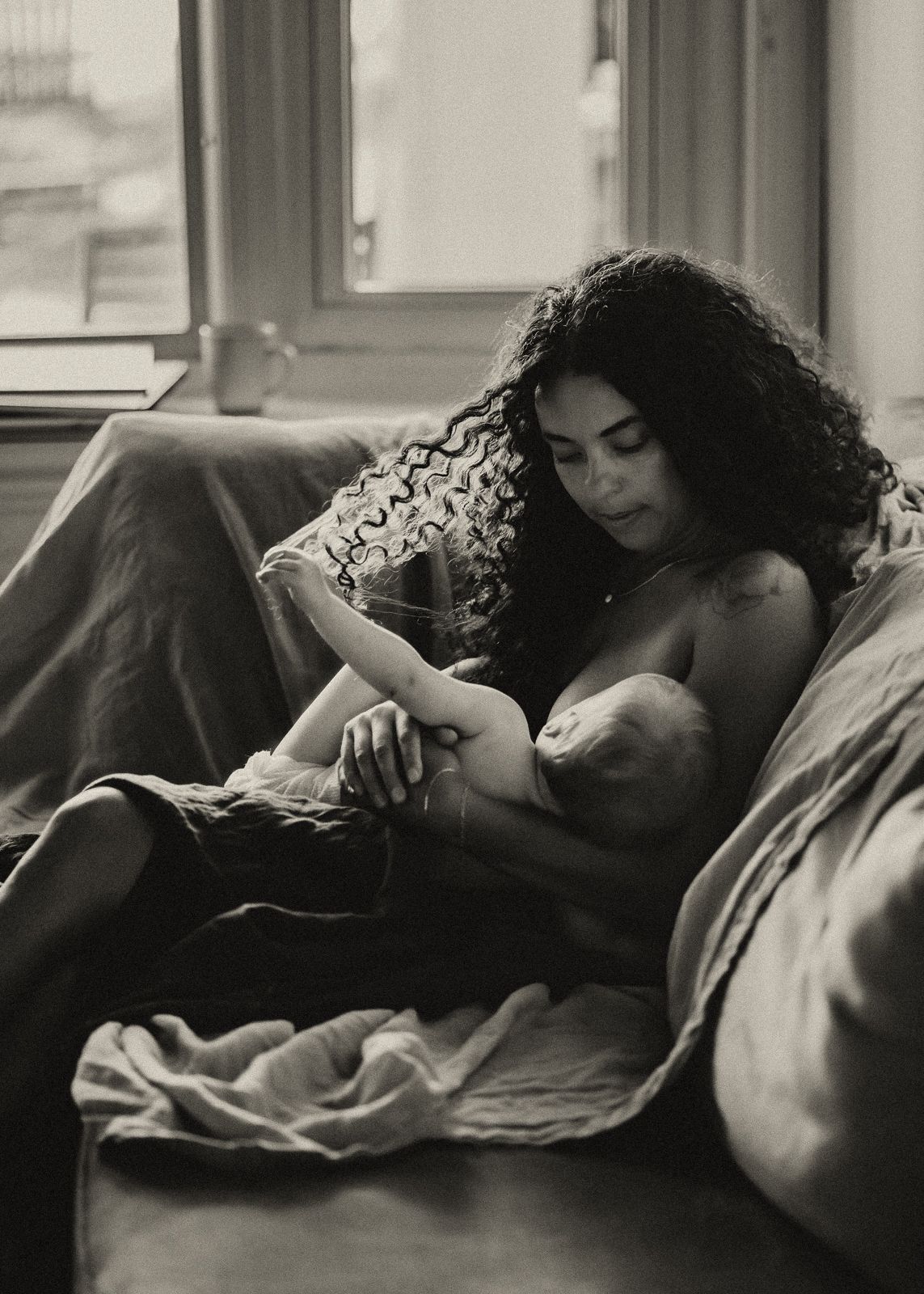 Black and white photograph of a tender breastfeeding moment in natural window light on a cozy bed.