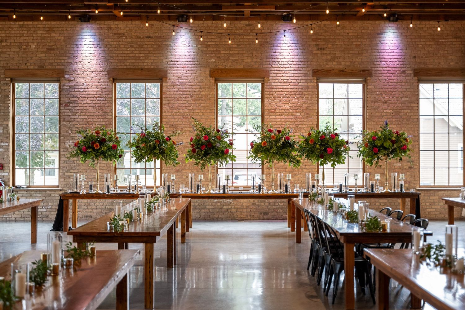 Elegant wedding reception venue with exposed brick walls, hanging plants, and long wooden tables illuminated by spotlights.