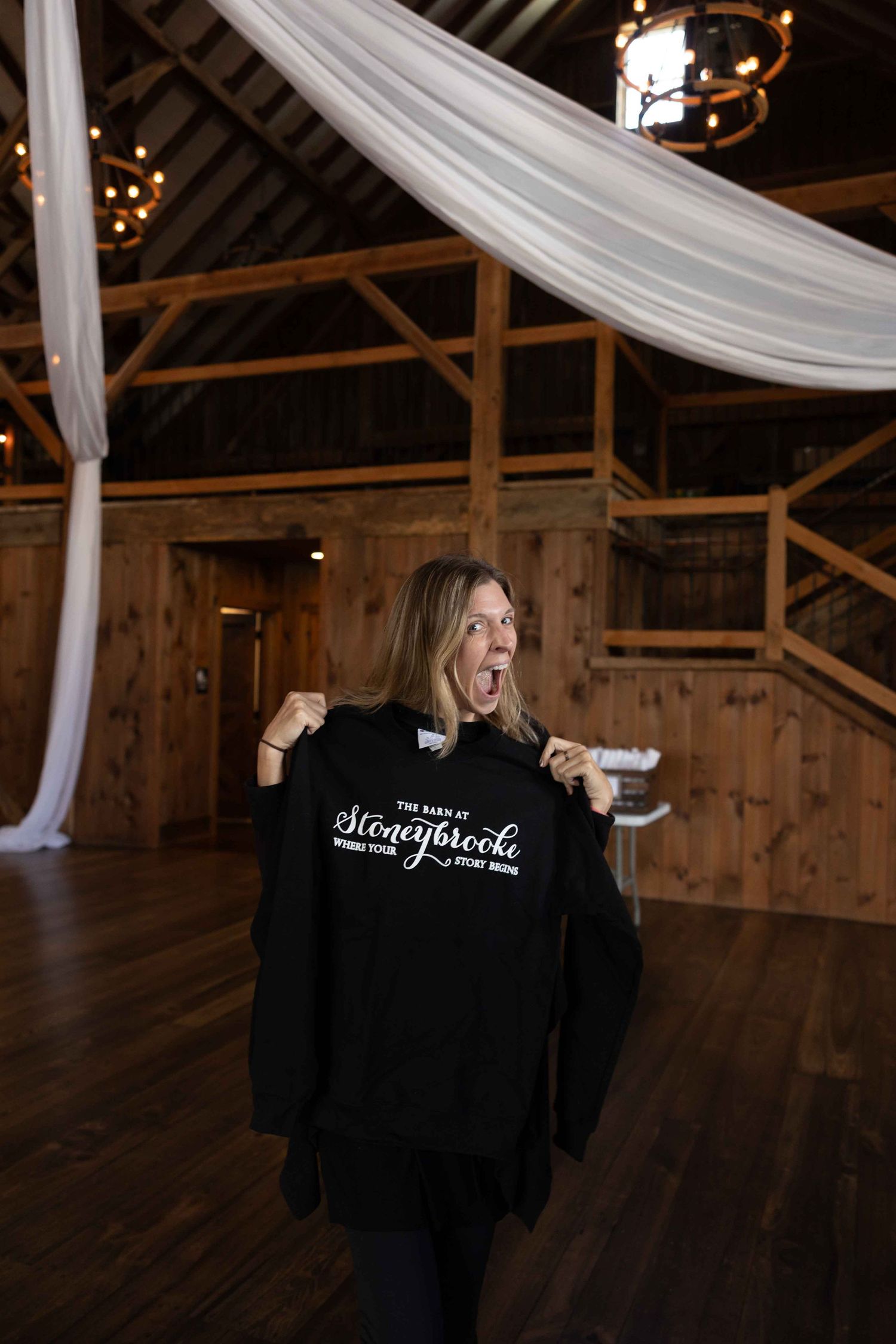 Someone holds up a black sweatshirt with text inside a rustic barn venue with white draping and wooden beams.