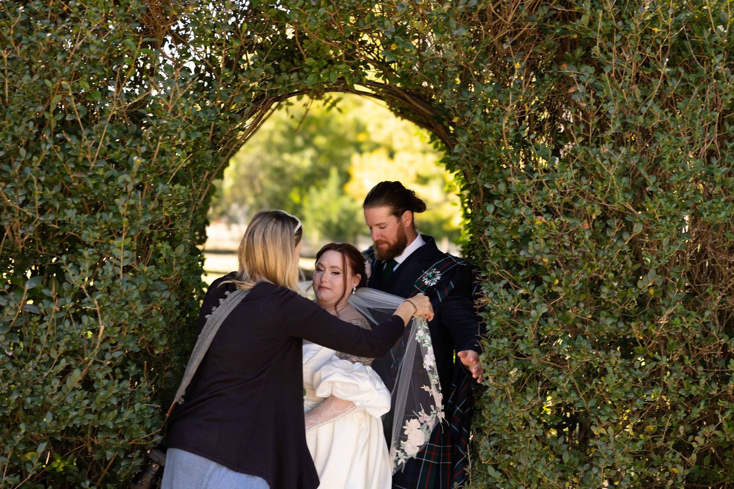 Three people share a warm embrace under a garden archway framed by lush green foliage on a sunny day.
