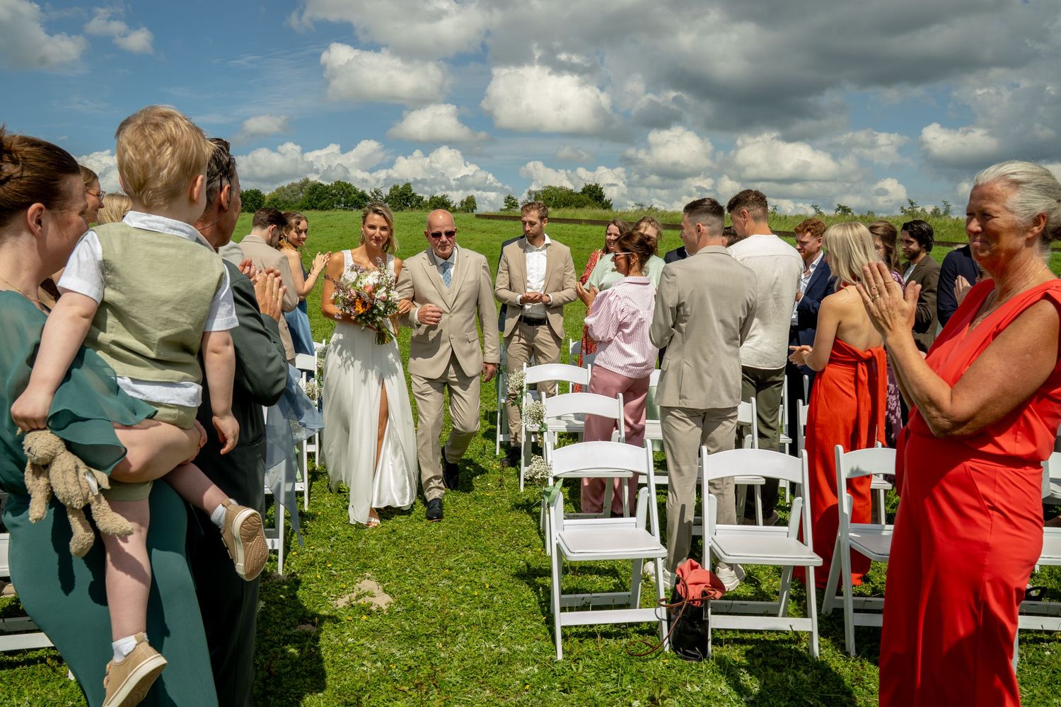Bride and groom walk down outdoor aisle during wedding ceremony surrounded by guests on a sunny day.