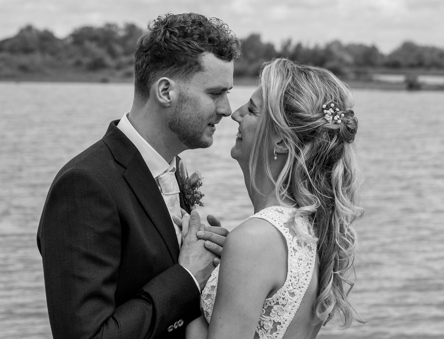 Black and white wedding photo of bride and groom sharing romantic moment by a lake.
